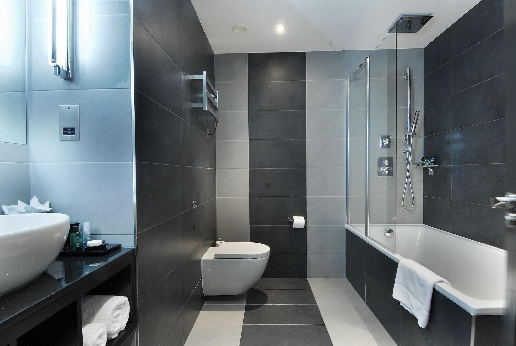 Montcalm Brewery Townhouse Hotel Londen Buitenkant foto The photo shows a modern bathroom featuring a sleek design. The walls are tiled in a combination of dark gray and light gray, creating a contemporary look. On the left, there is a countertop with a basin and some toiletries placed on it. The middle s