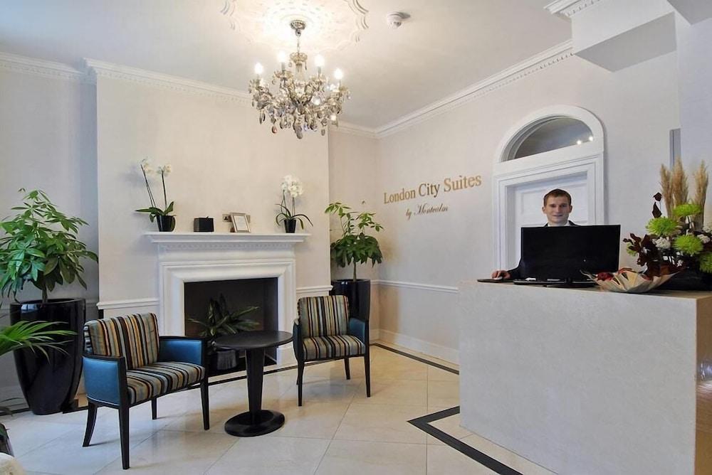 Montcalm Brewery Townhouse Hotel Londen Buitenkant foto The photo shows the interior of a reception area in a hotel called "London City Suites." The space features a light-colored wall with the hotel's name displayed. There is a stylish chandelier hanging from the ceiling, providing warm lighting. 

In th