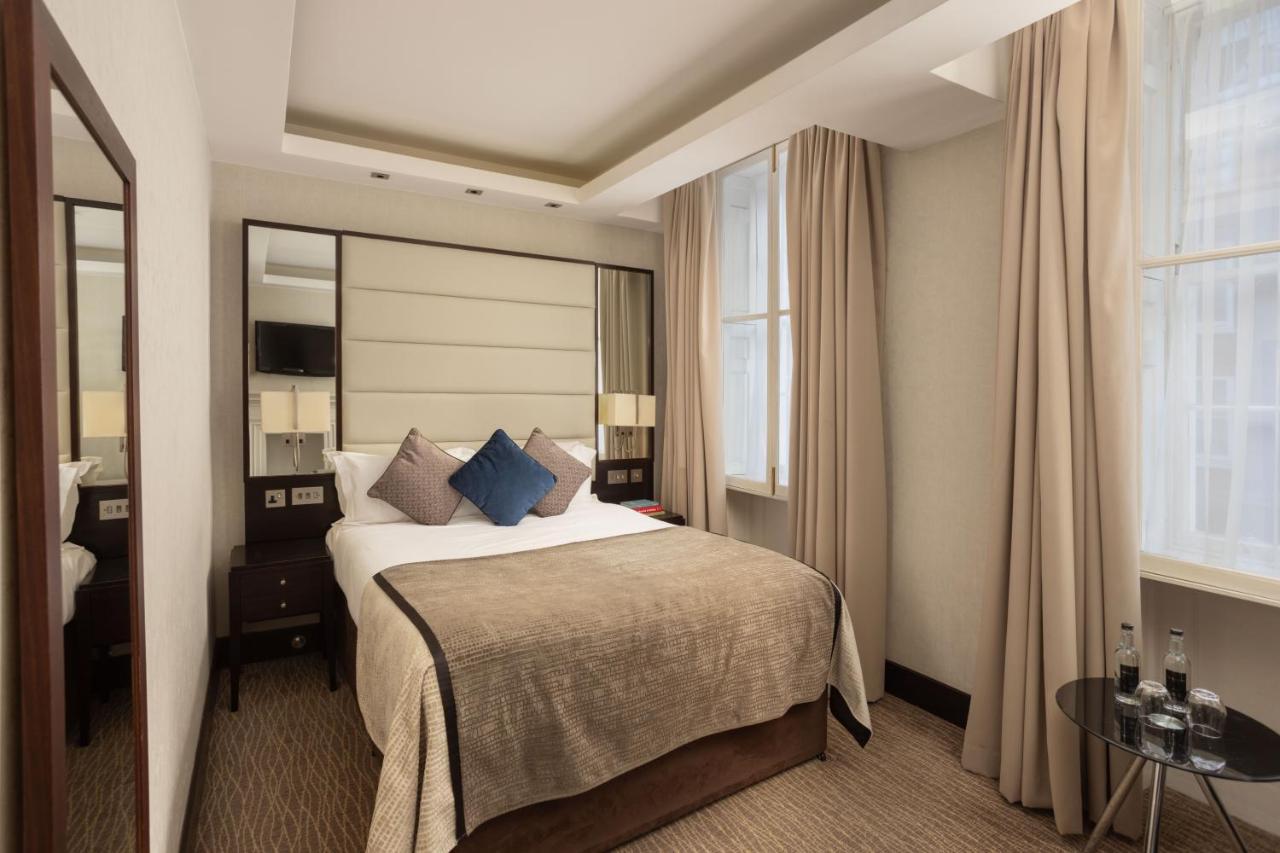 Montcalm Brewery Townhouse Hotel Londen Buitenkant foto The photo shows a cozy hotel bedroom. The room features a neatly made double bed with a beige bedspread and decorative pillows, including a blue one. There are bedside tables on either side of the bed with lamps. The walls are painted in a soft color