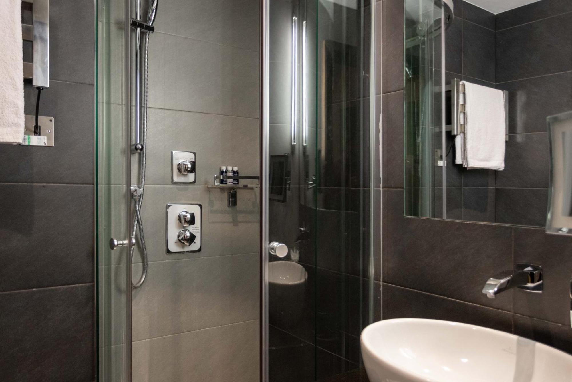 Montcalm Brewery Townhouse Hotel Londen Buitenkant foto The photo shows a modern bathroom with a glass shower enclosure. The shower features a handheld showerhead and various controls mounted on the wall. The walls are tiled in dark, sleek tones, contributing to a contemporary aesthetic. There is a round 