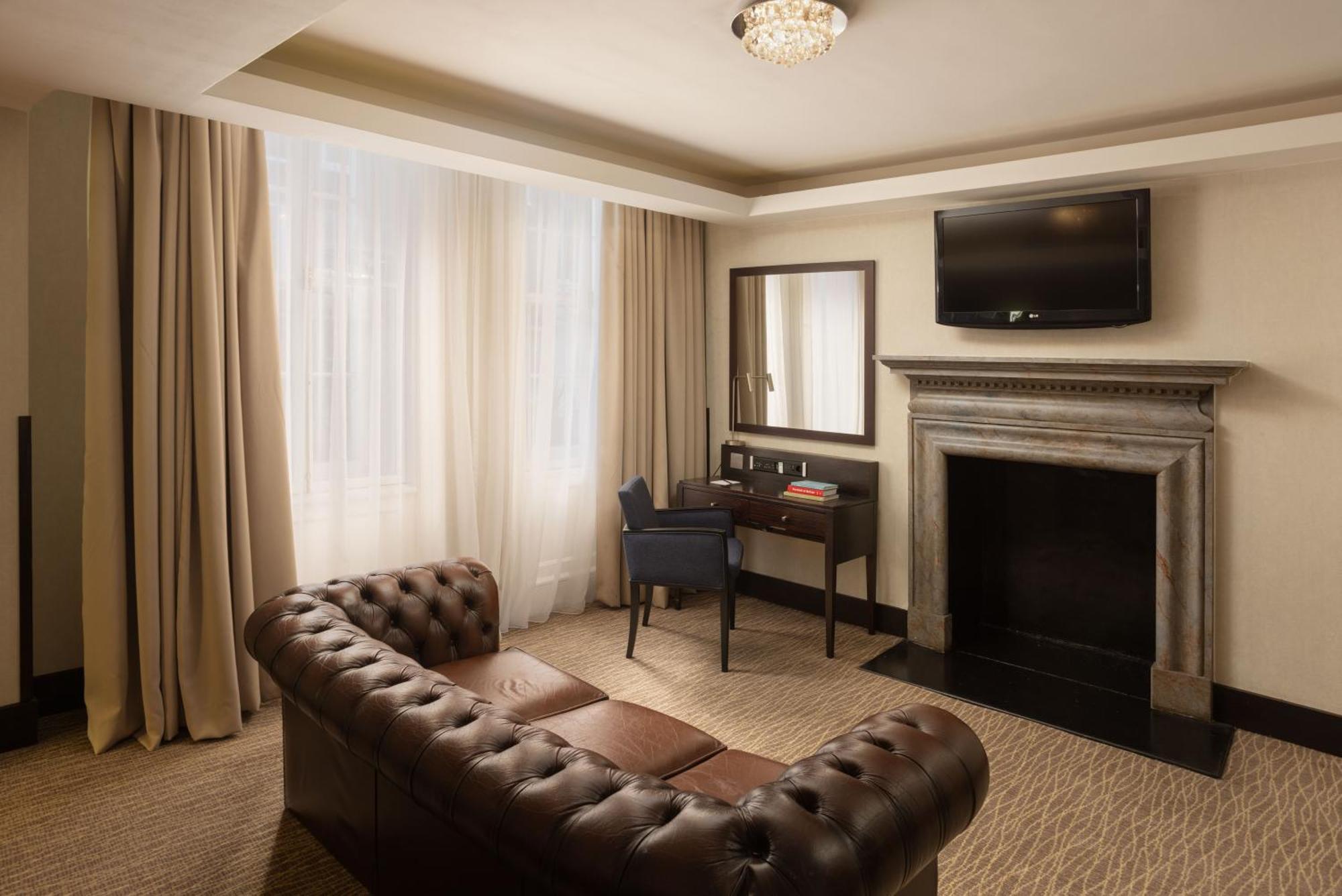 Montcalm Brewery Townhouse Hotel Londen Buitenkant foto The photo shows a stylish interior of a room, likely a living area or a hotel suite. It features a dark leather chesterfield sofa with tufted upholstery. Opposite the sofa is a small wooden desk with a chair, positioned near a wall-mounted flat-scree