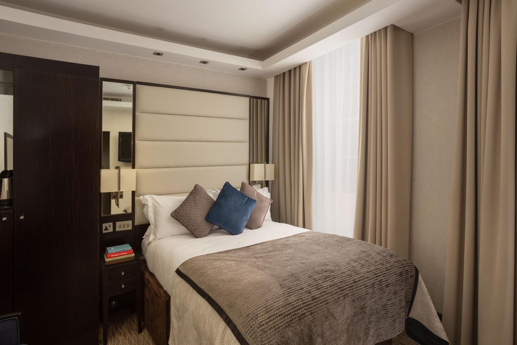 Montcalm Brewery Townhouse Hotel Londen Buitenkant foto The photo shows a modern hotel room with a cozy design. It features a double bed with a light-colored headboard and a soft, stylish blanket. There are decorative pillows on the bed, including a blue one. On either side of the bed, there are small nig