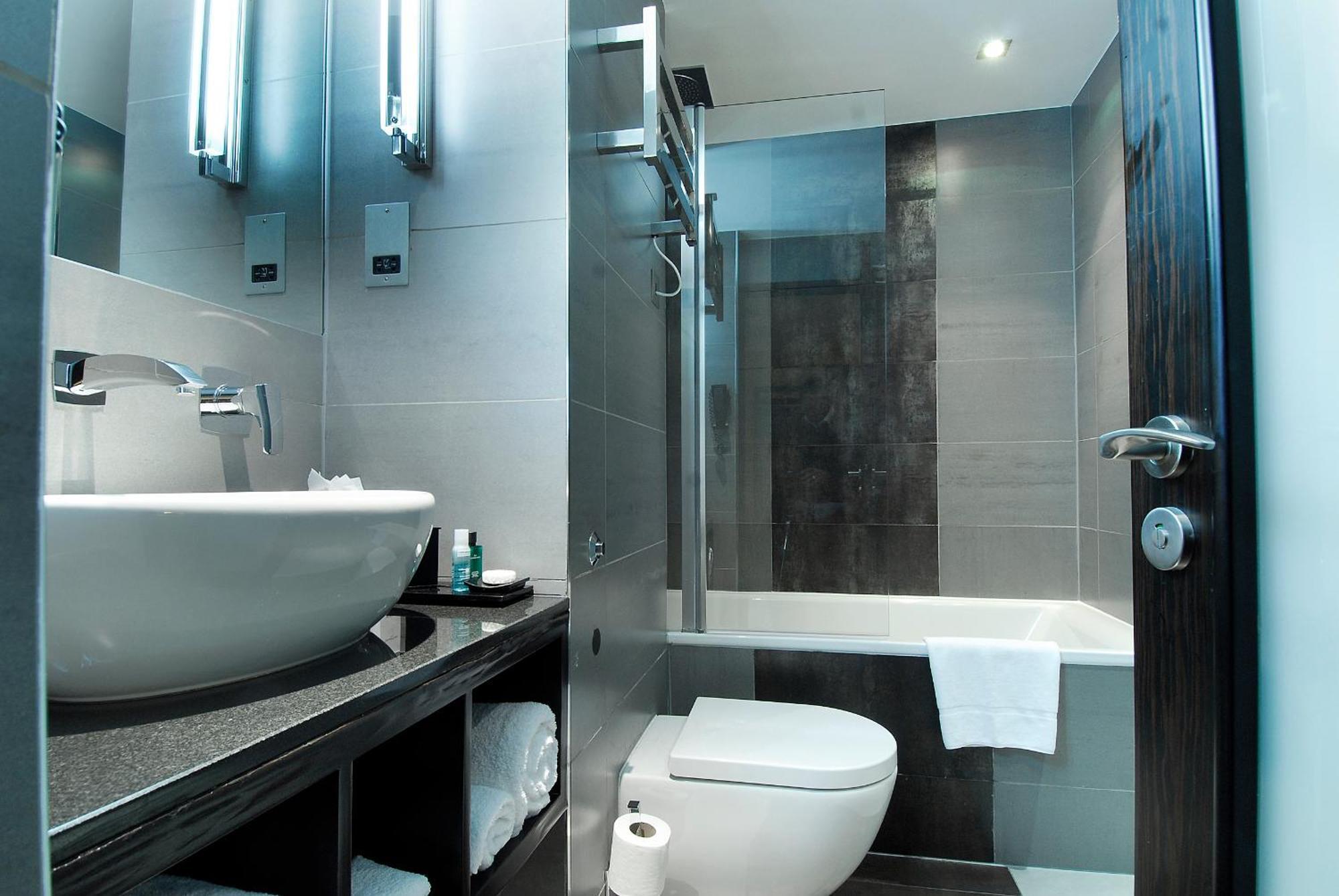 Montcalm Brewery Townhouse Hotel Londen Buitenkant foto The photo shows a modern bathroom with sleek design elements. It features a curved white sink on a dark countertop, with a mirror above that is illuminated by stylish lighting fixtures. Next to the sink, there's a toilet, and on the opposite side, a 