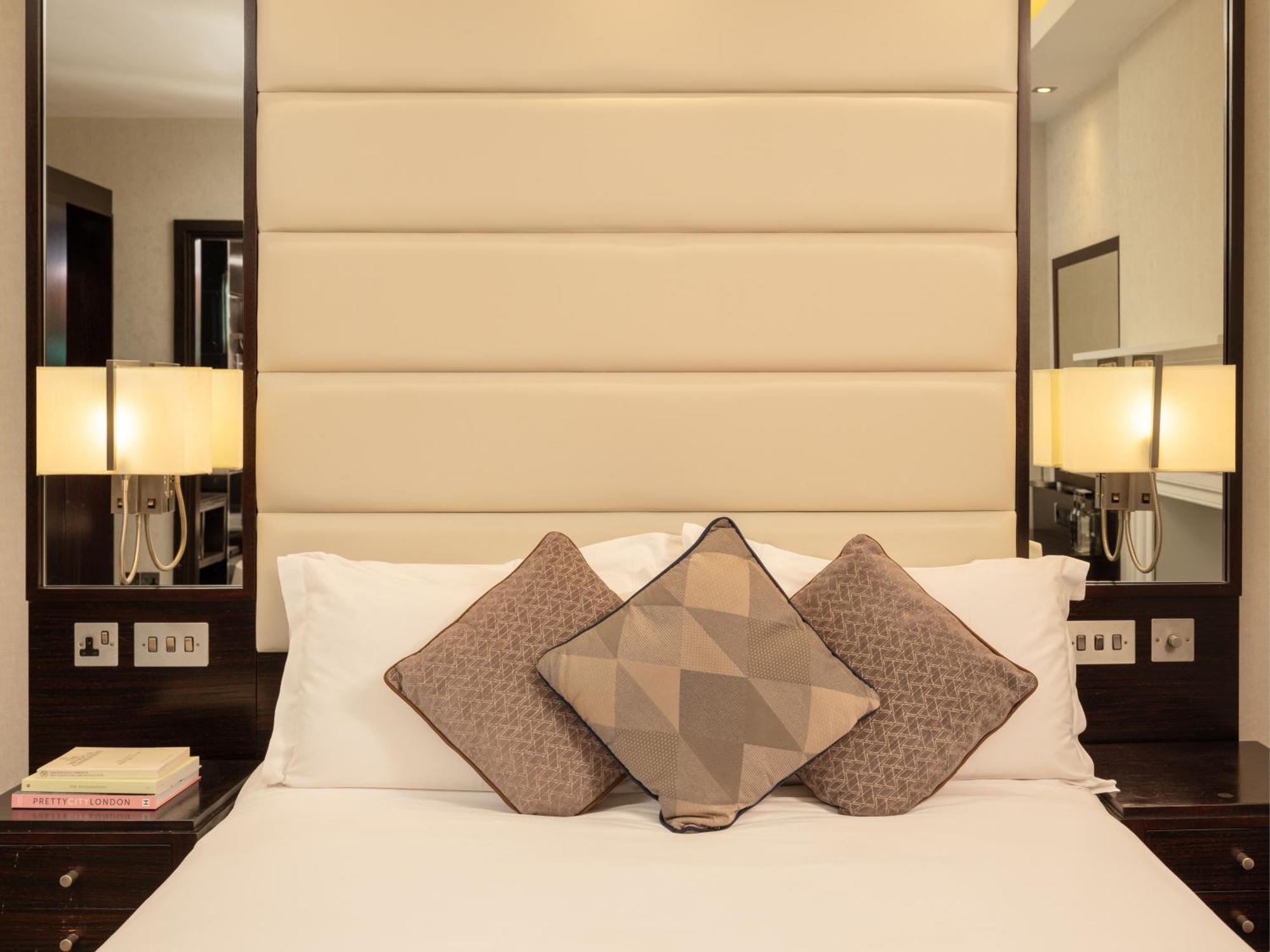 Montcalm Brewery Townhouse Hotel Londen Buitenkant foto The image shows a cozy bedroom setting. The bed features a headboard upholstered in a light beige fabric. It is adorned with three decorative pillows in varying patterns and colors, placed on white bedding. On either side of the bed, there are modern