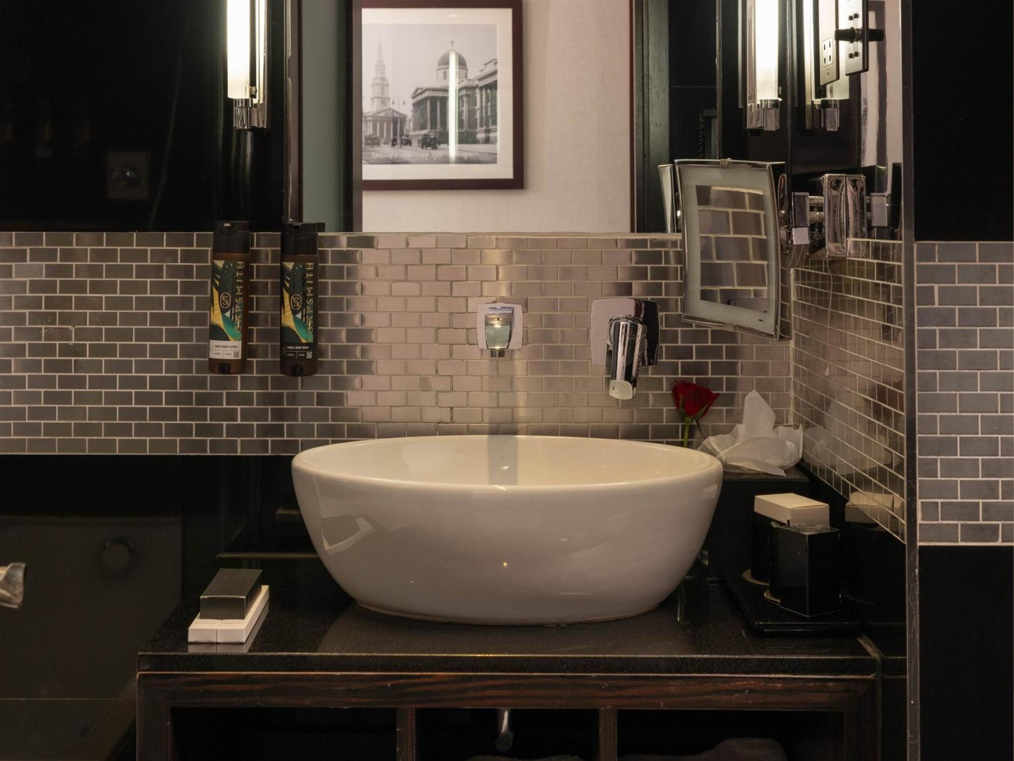 Montcalm Brewery Townhouse Hotel Londen Buitenkant foto The photo shows a modern bathroom with a stylish design. It features a large, round white sink placed on a dark wooden vanity. The walls are adorned with a mix of gray and silver tile, creating a sleek and contemporary look. Above the sink, there is 