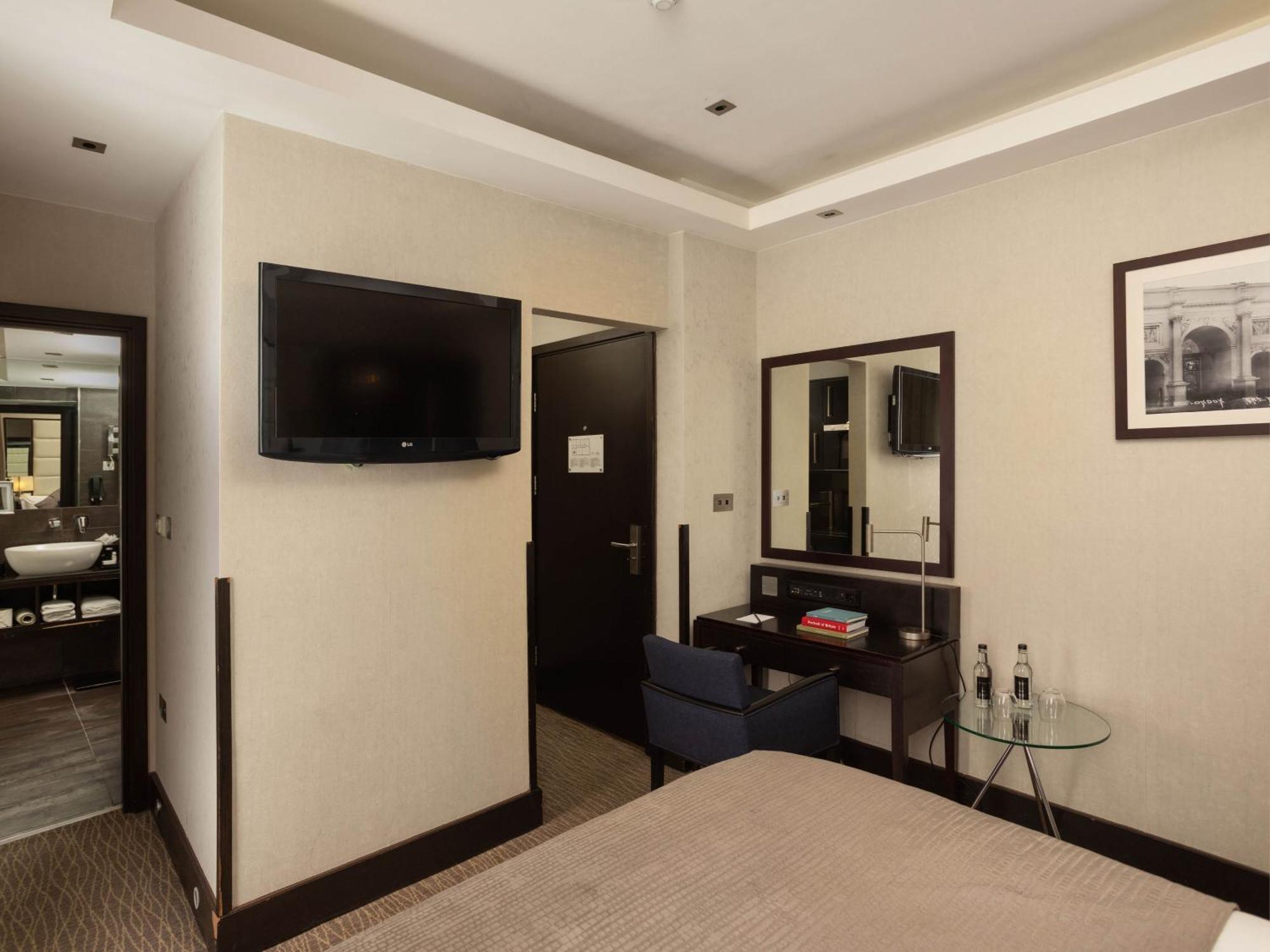 Montcalm Brewery Townhouse Hotel Londen Buitenkant foto The photo shows a hotel room with a modern and minimalist design. On the left, there's a wall-mounted flat-screen TV. In the center, there's a bed with a neutral-colored bedspread. To the right of the bed, there is a small desk with a mirror above it