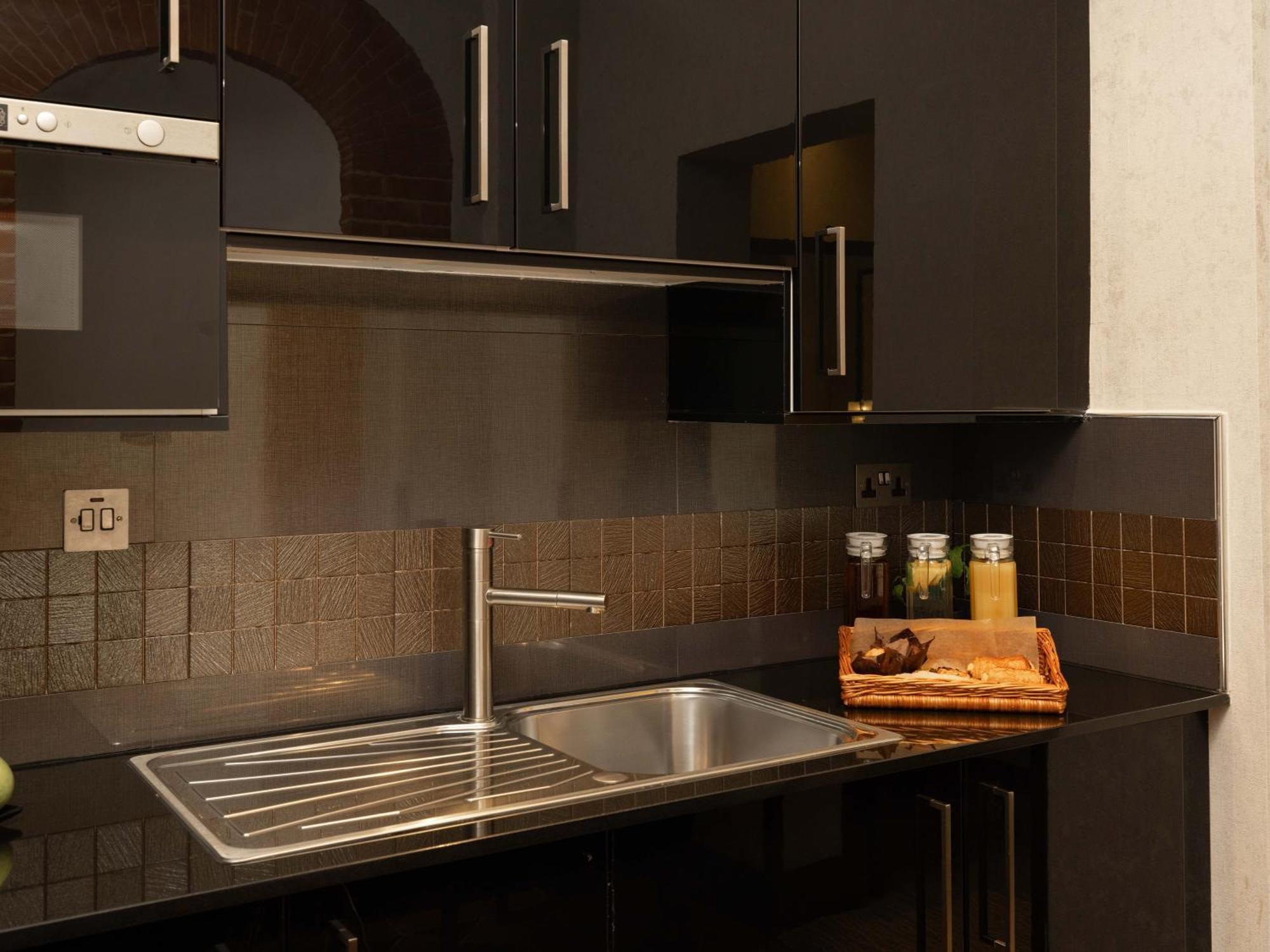 Montcalm Brewery Townhouse Hotel Londen Buitenkant foto The photo shows a modern kitchen with a sleek design. It features dark cabinetry with a glossy finish and stainless steel handles. There is a stainless steel sink with a simple faucet, and a countertop made of black material. Above the sink, there ar