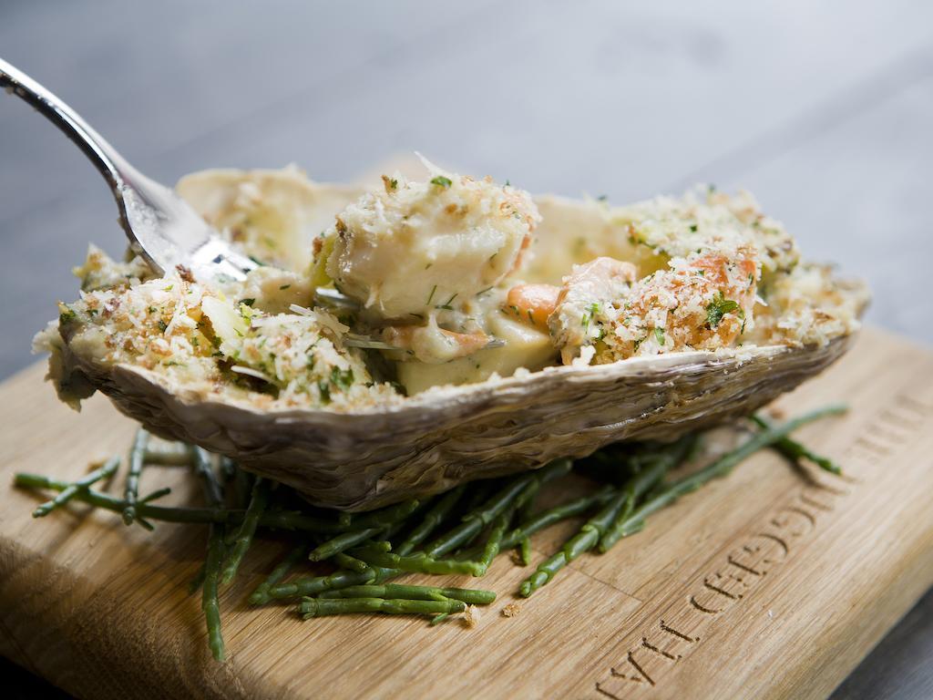 Montcalm Brewery Townhouse Hotel Londen Buitenkant foto The photo shows a large oyster shell filled with a mixture that appears to include seafood, herbs, and breadcrumbs. The dish is presented on a wooden board with some greenery, possibly a type of seaweed or garnishing. A fork is placed beside the oyst