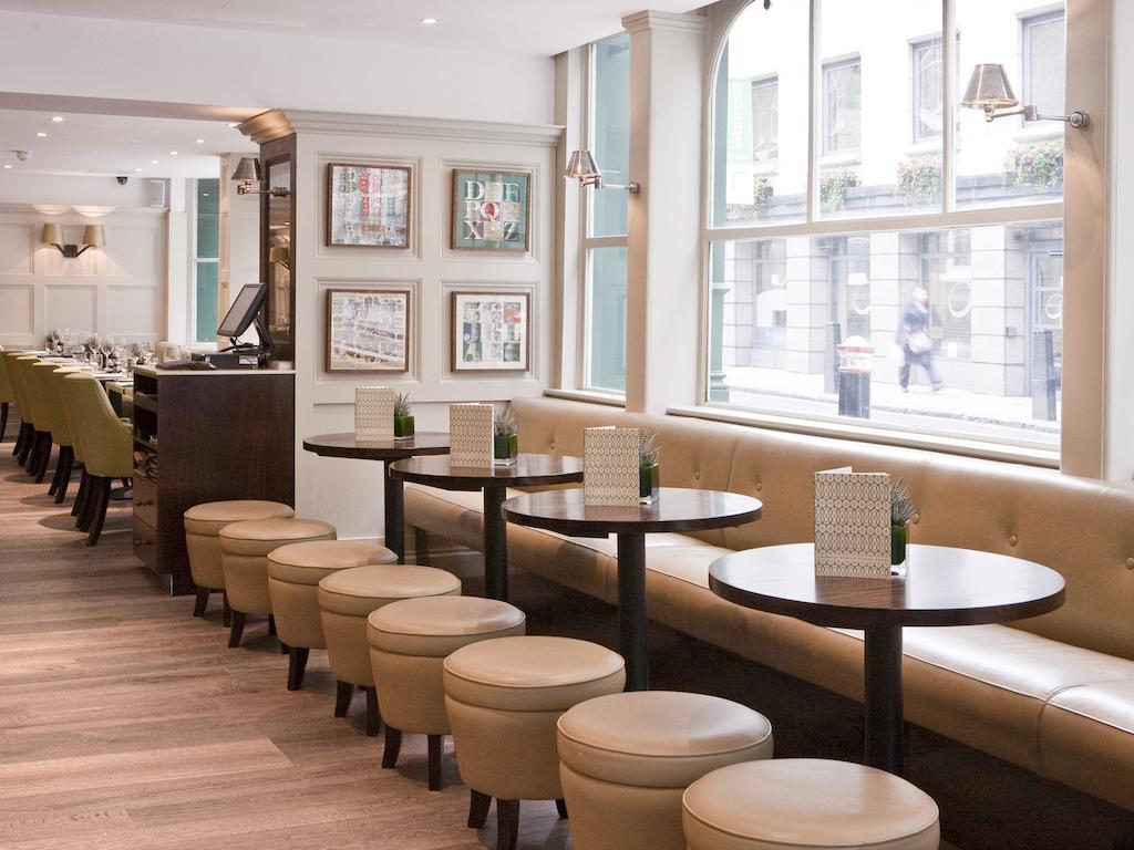 Montcalm Brewery Townhouse Hotel Londen Buitenkant foto The photo displays a modern café or restaurant interior. There is a cozy seating area featuring light-colored, upholstered benches along the wall, accompanied by small round tables. The tables are paired with circular stools. The decor includes frame