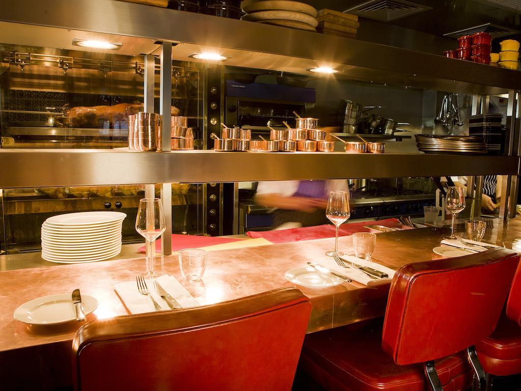 Montcalm Brewery Townhouse Hotel Londen Buitenkant foto The photo shows a restaurant interior featuring a bar or counter area. There are red chairs arranged around a copper-topped table where dining utensils and plates are set. Above the counter, there are shelves displaying various cooking pots and kitch