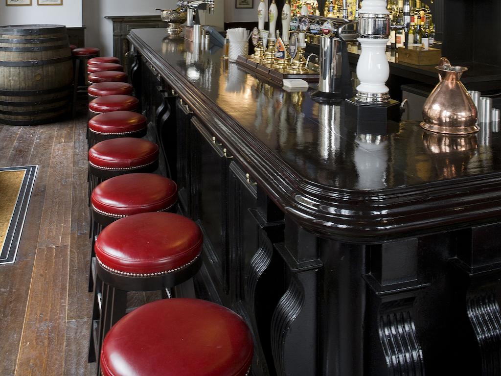 Montcalm Brewery Townhouse Hotel Londen Buitenkant foto The photo shows a bar setting with a long, polished dark wooden bar counter. Attached to the counter are several bar stools with red leather seats. Behind the bar, there are various bottles, suggesting a well-stocked bar. The flooring appears to be w
